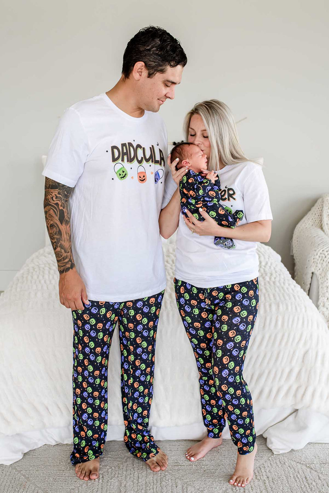 Father and mother standing beside bed wearing matching 'DADCULA' and 'MOMSTER' Halloween shirts with candy-print pajama pants, mother kissing newborn's head