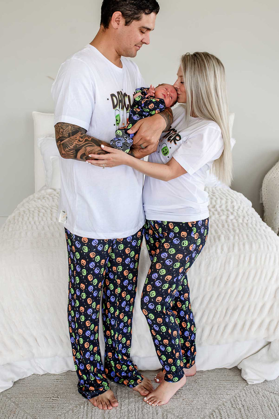 Father and mother standing beside bed wearing matching Halloween Candy Crawl pajamas, mother kissing newborn's head, both in candy-print pajama pants