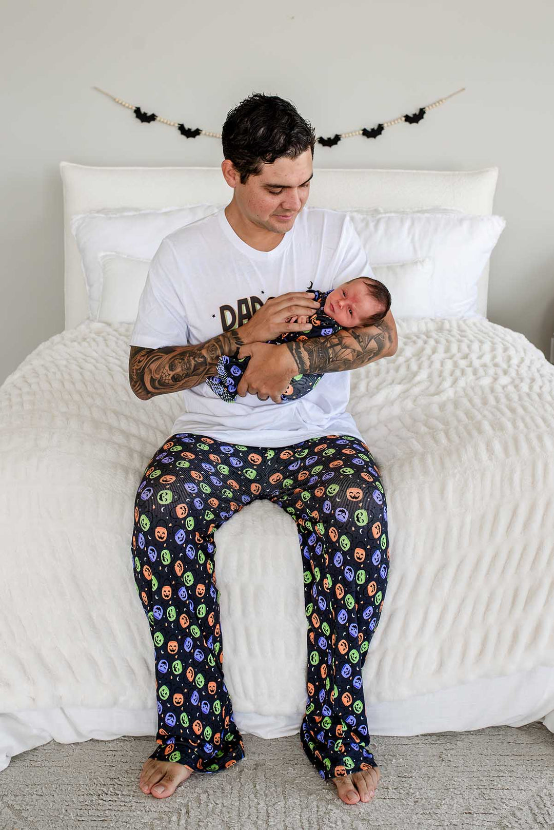 Father sitting on bed in 'DADCULA' t-shirt and candy-print pajama pants, holding a newborn baby, Halloween bat garland on the wall behind him