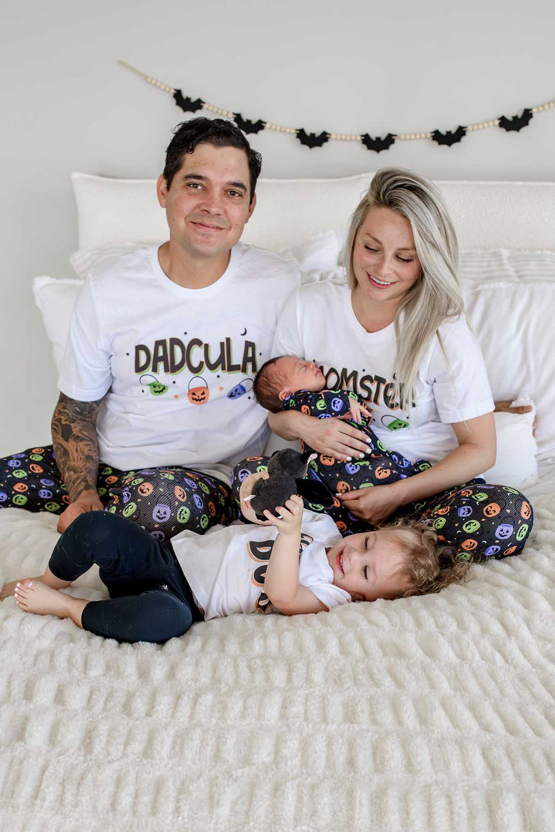 Family on a bed wearing matching Halloween Candy Crawl pajamas: father in 'DADCULA' shirt, mother in 'MOMSTER' shirt holding newborn, toddler playing with a small dog, bat garland on wall behind them
