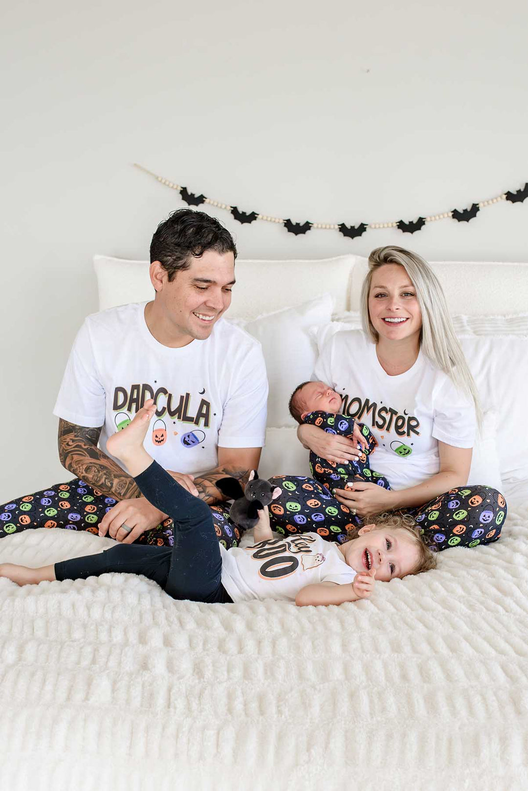 Family on a bed wearing matching Halloween Candy Crawl pajamas: father in 'DADCULA' shirt, mother in 'MOMSTER' shirt holding newborn, toddler in matching tee playing with a small dog, bat garland on wall behind them
