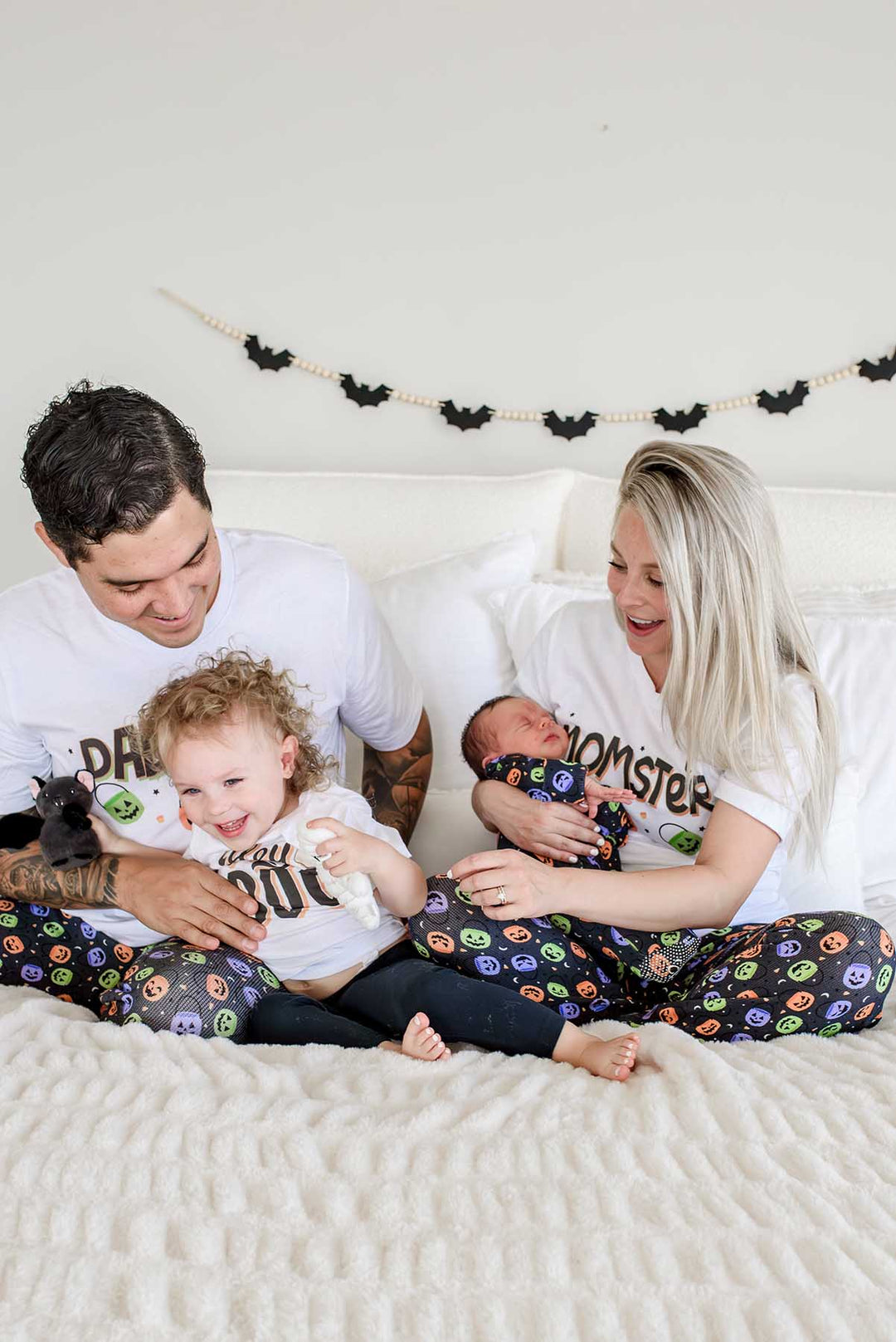 Family on a bed wearing matching Halloween Candy Crawl pajamas: father in 'DADCULA' shirt with toddler, mother in 'MOMSTER' shirt holding newborn, small dog beside them, bat garland on wall behind