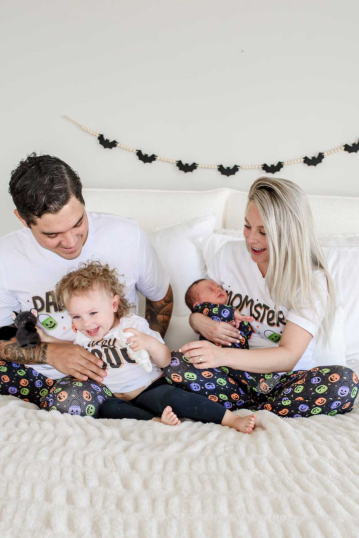 Family on a bed wearing matching Halloween Candy Crawl pajamas: father in 'DADCULA' shirt with toddler, mother in 'MOMSTER' shirt holding newborn, small dog beside them, bat garland on wall behind