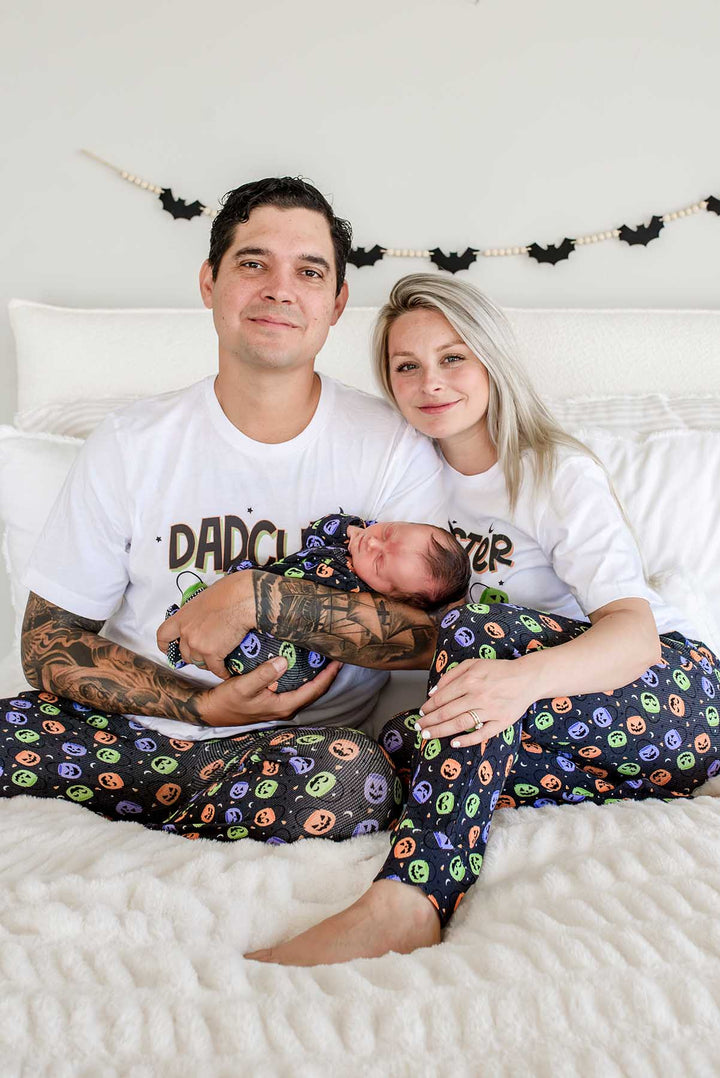 Father and mother sitting on bed wearing matching 'DADCULA' and 'MOMSTER' shirts and candy-print pajama pants, holding a newborn, bat garland on wall behind them