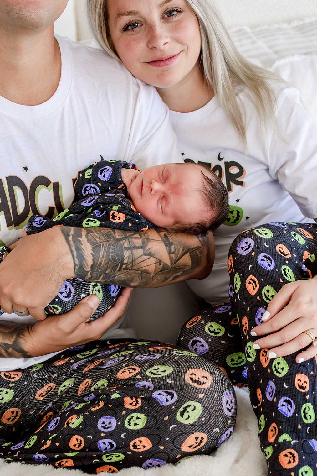 Close-up of parents in matching 'DADCULA' and 'MOMSTER' shirts holding a sleeping newborn wrapped in candy-print fabric