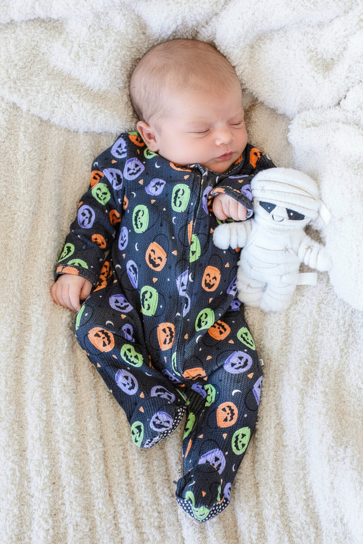 Baby lying on a fluffy blanket in a bamboo waffle zipper footie with colorful jack-o-lantern print, beside a mummy plush toy