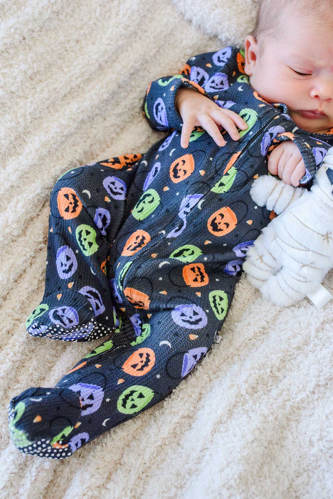Sleeping baby lying on a fluffy blanket in a bamboo waffle zipper footie with colorful jack-o-lantern print, beside a mummy plush toy