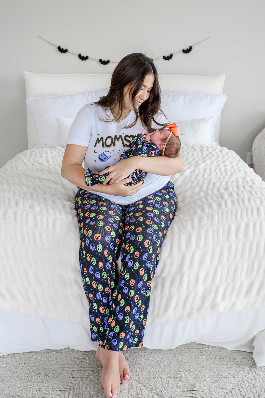 Mother sitting on bed wearing 'MOMSTER' shirt and candy-print pajama pants, holding a newborn in an orange bow, bat garland on wall behind her