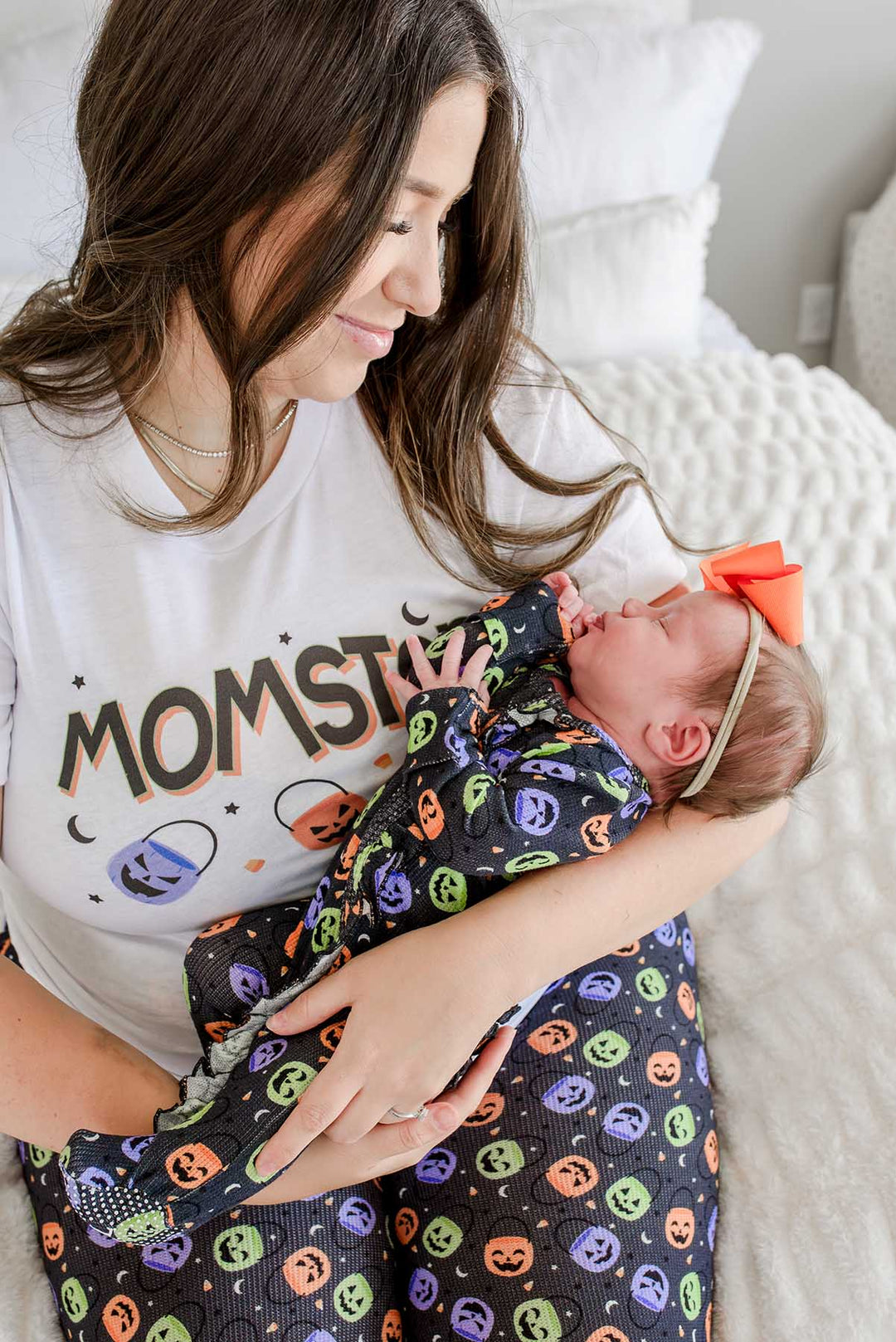 Mother in 'MOMSTER' shirt holding a newborn wearing an orange bow, wrapped in a bamboo waffle ruffle footie with colorful jack-o-lantern print
