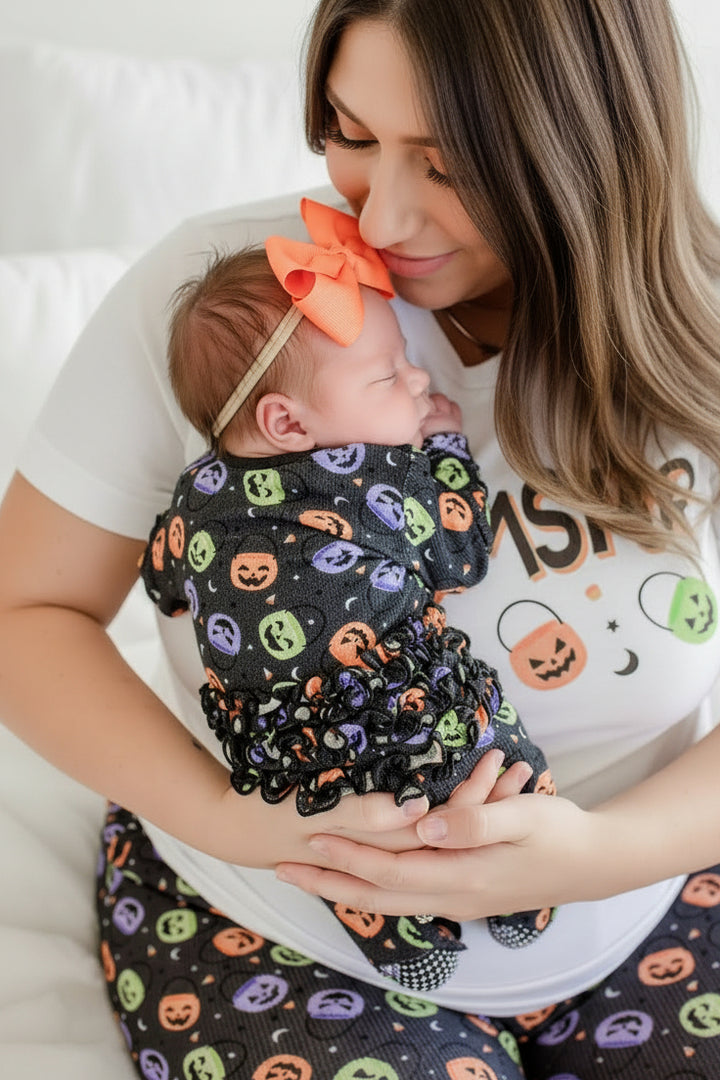 Mother in white 'MOMSTER' shirt holding a newborn wearing an orange bow, wrapped in a bamboo waffle ruffle footie with colorful jack-o-lantern print