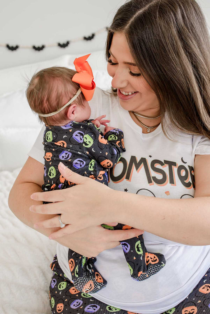 Mother in 'MOMSTER' shirt smiling at a baby with an orange bow wearing a bamboo waffle ruffle footie with colorful jack-o-lantern print