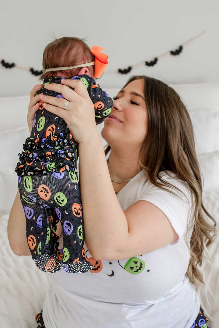 Mother lifting a baby wearing an orange bow and a bamboo waffle ruffle footie with colorful jack-o-lantern print, bat garland on wall behind