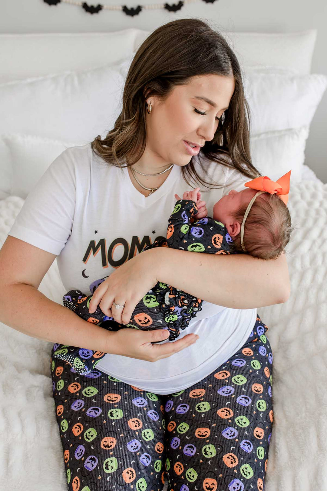 Mother in 'MOMSTER' Halloween shirt and candy-print pajama pants holding a newborn wearing an orange bow, swaddled in matching print