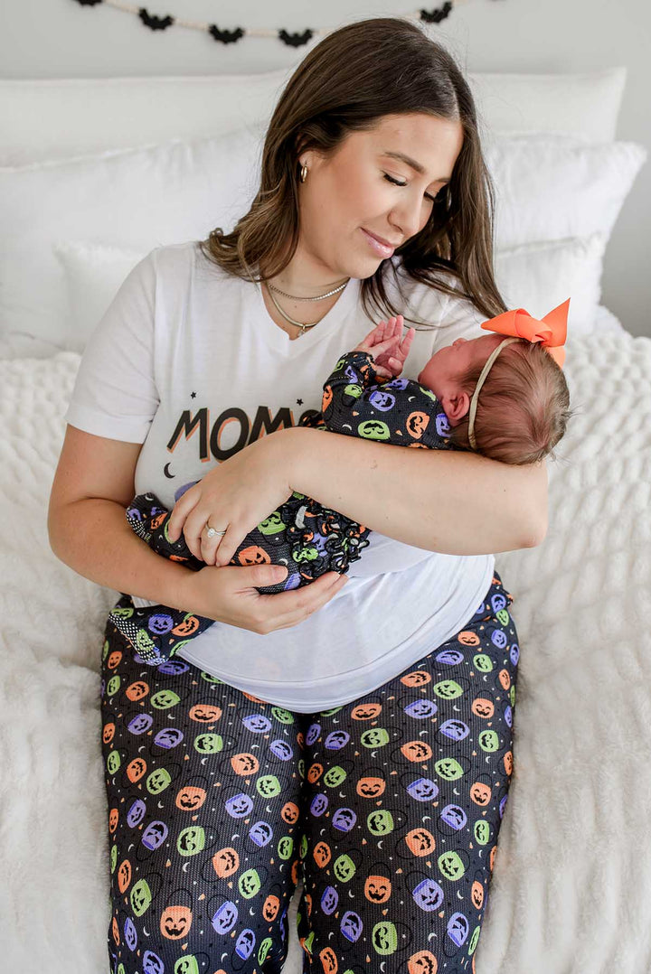 Mother in 'MOMSTER' shirt and candy-print pajama pants holding a newborn with an orange bow, bat garland on wall behind her