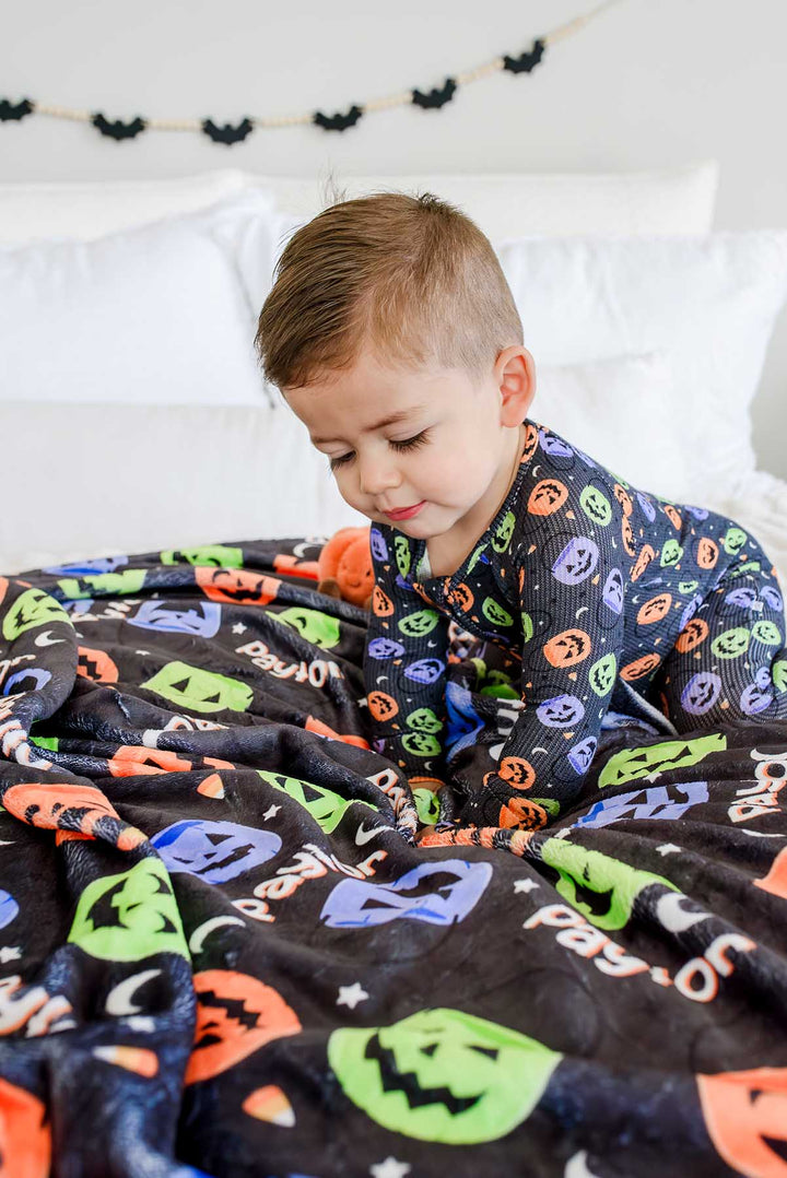 Toddler sitting on a bed wrapped in a personalized Halloween blanket with colorful jack-o-lantern print and name text, bat garland on wall behind