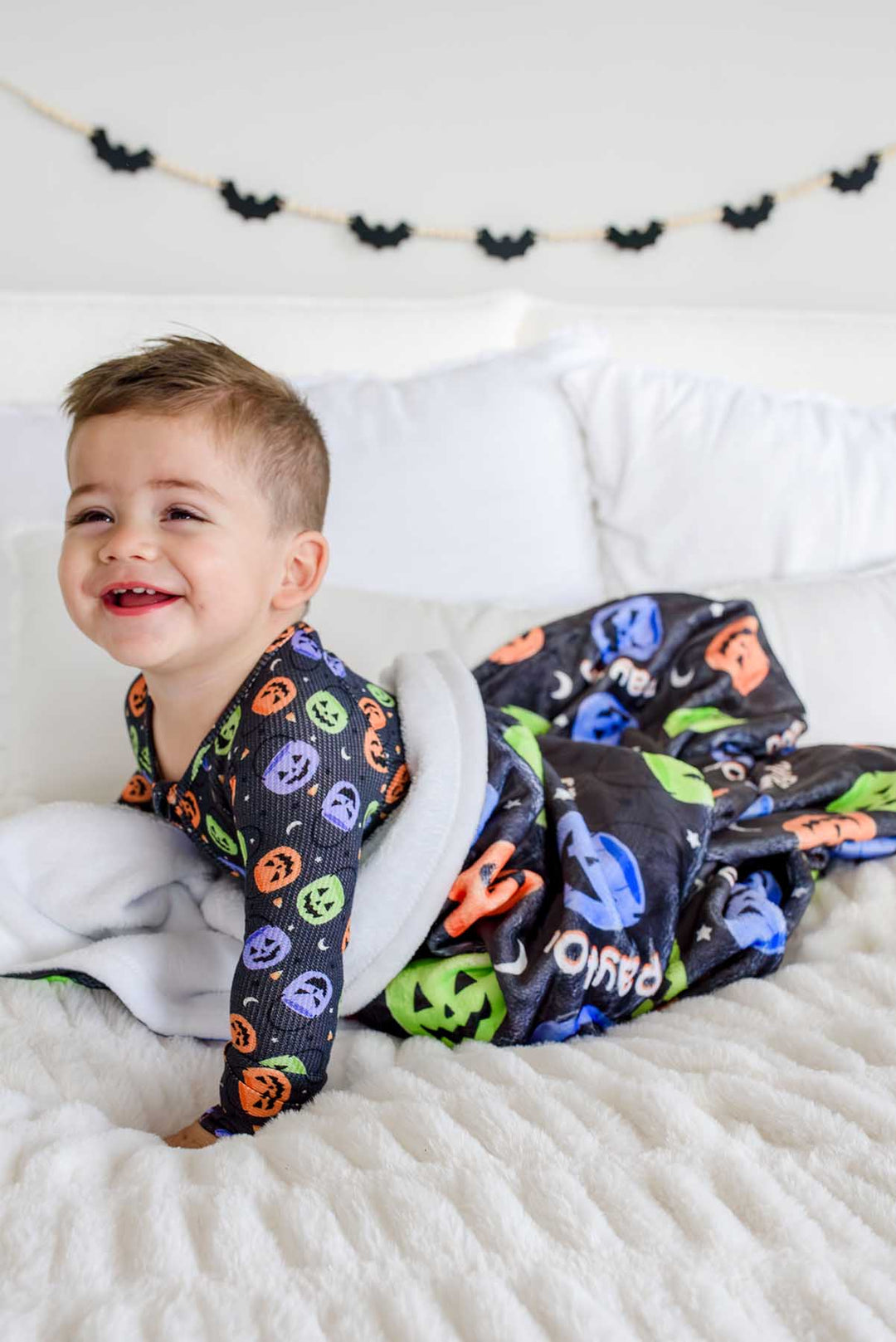 Baby crawling on a bed on top of a personalized Halloween blanket with colorful jack-o-lantern print and name text, bat garland on wall behind