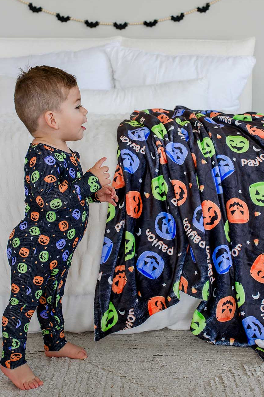 Toddler in candy-print pajamas standing beside a bed draped with a personalized Halloween blanket featuring jack-o-lantern print and name text, bat garland on wall behind