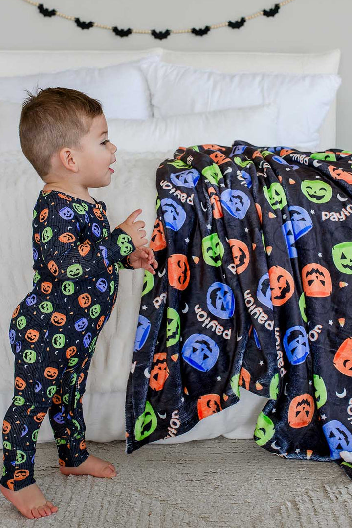 Toddler in candy-print pajamas standing beside a bed draped with a personalized Halloween blanket featuring jack-o-lantern print and name text, bat garland on wall behind