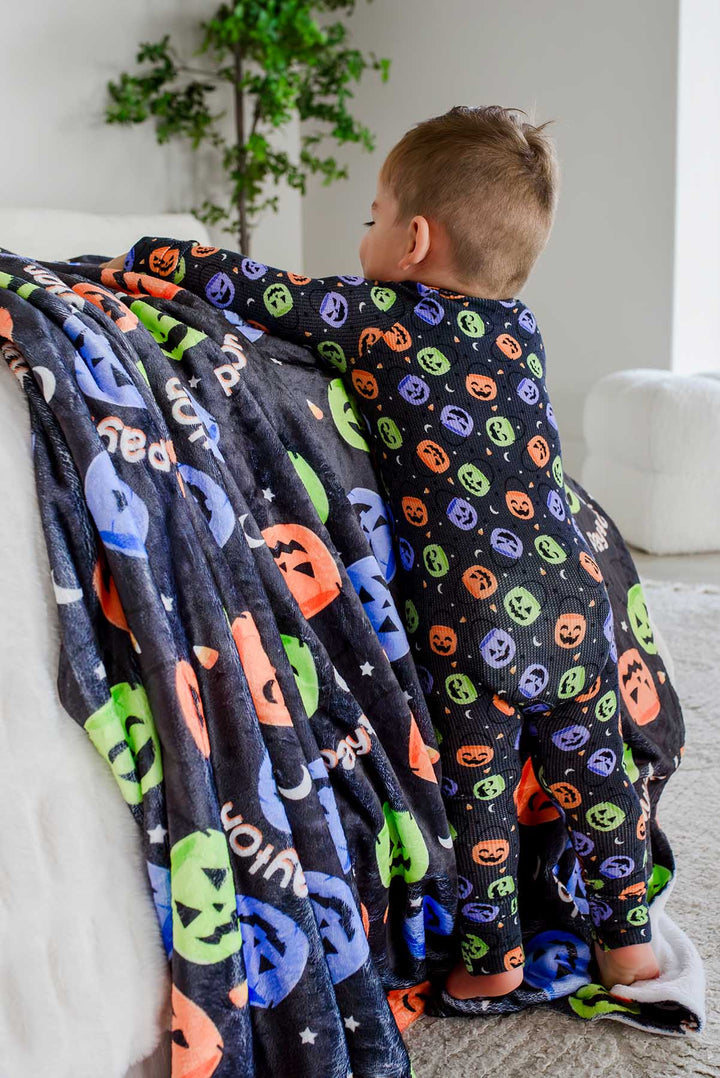 Toddler in candy-print pajamas climbing on a chair draped with a personalized Halloween blanket featuring jack-o-lantern print and name text