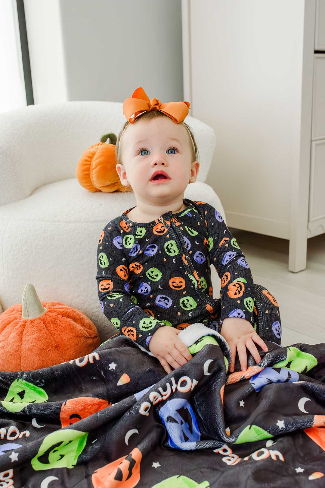 Baby with an orange bow sitting beside pumpkin decor, wrapped in a personalized Halloween blanket with colorful jack-o-lantern print and name text