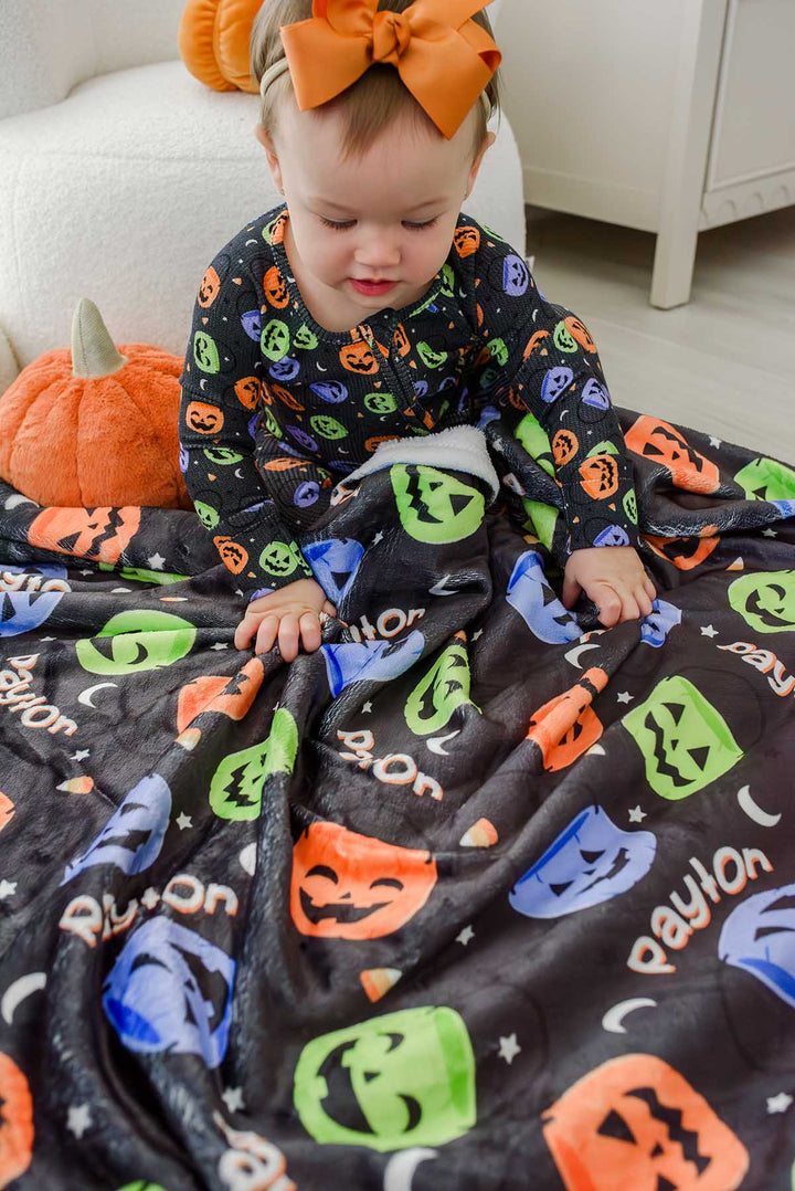 Baby with a large orange bow sitting beside pumpkin decor, wrapped in a personalized Halloween blanket with colorful jack-o-lantern print and repeating name text