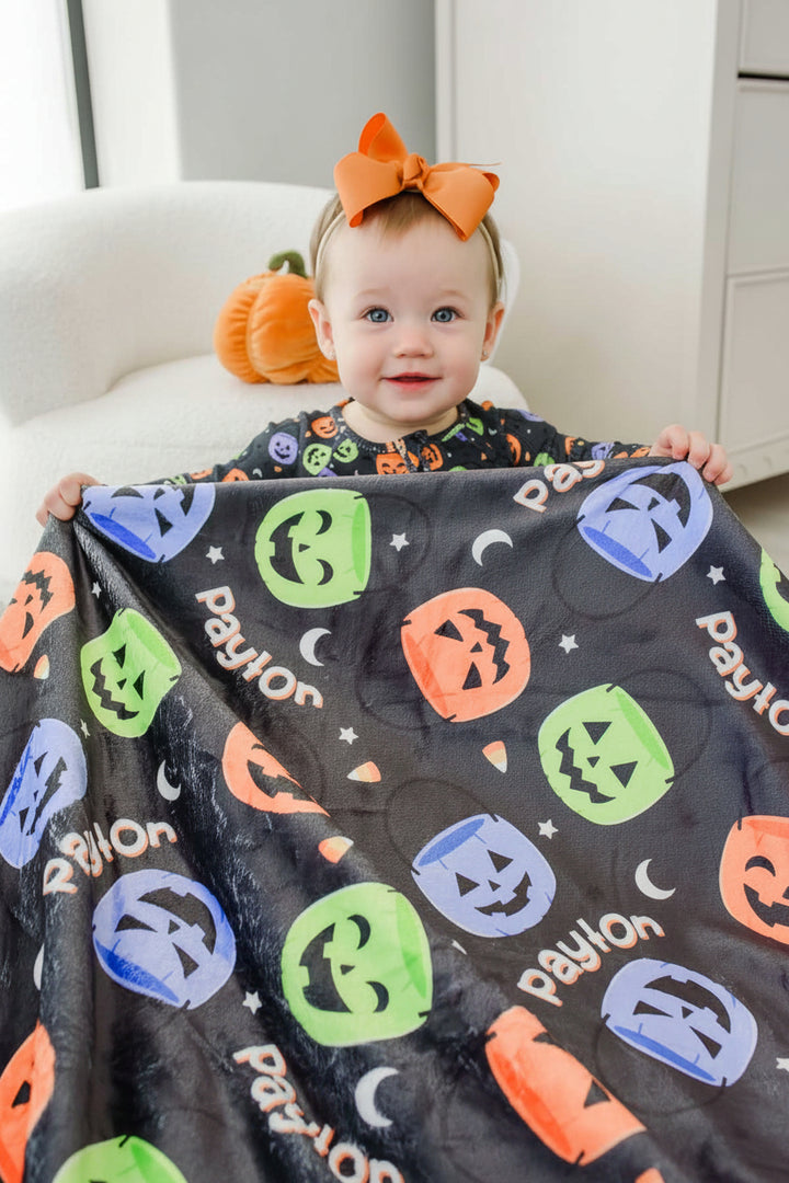 Baby with an orange bow holding up a personalized Halloween blanket with repeating name text and colorful jack-o-lantern print