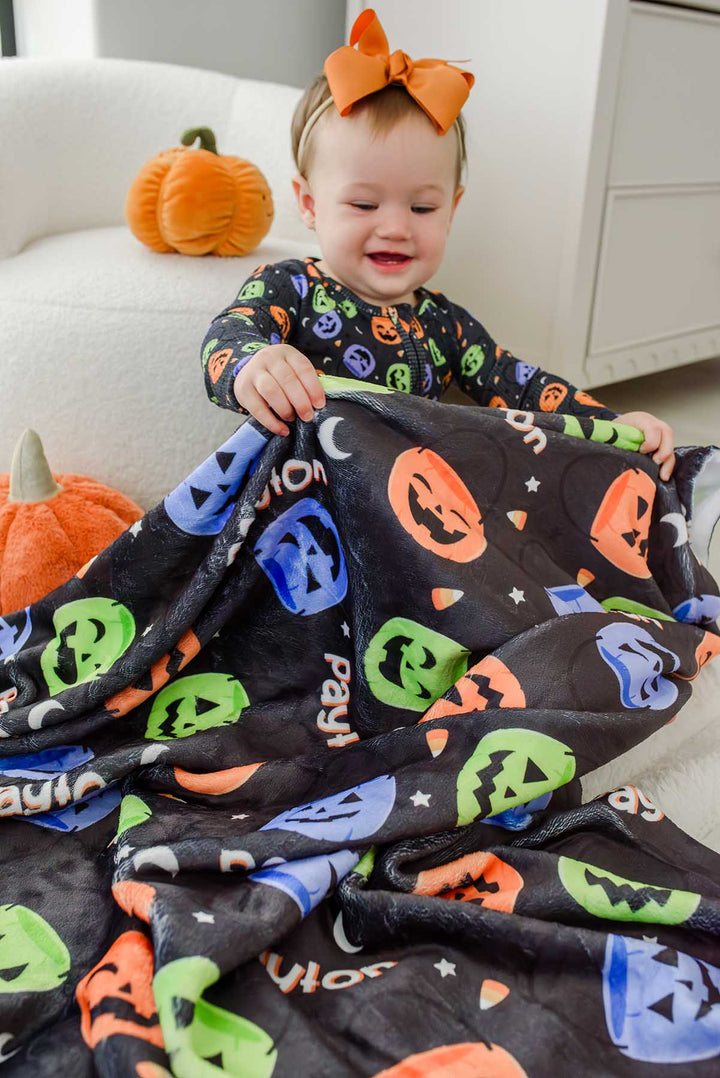 Baby with an orange bow playing with a personalized Halloween blanket featuring colorful jack-o-lantern print and repeating name text