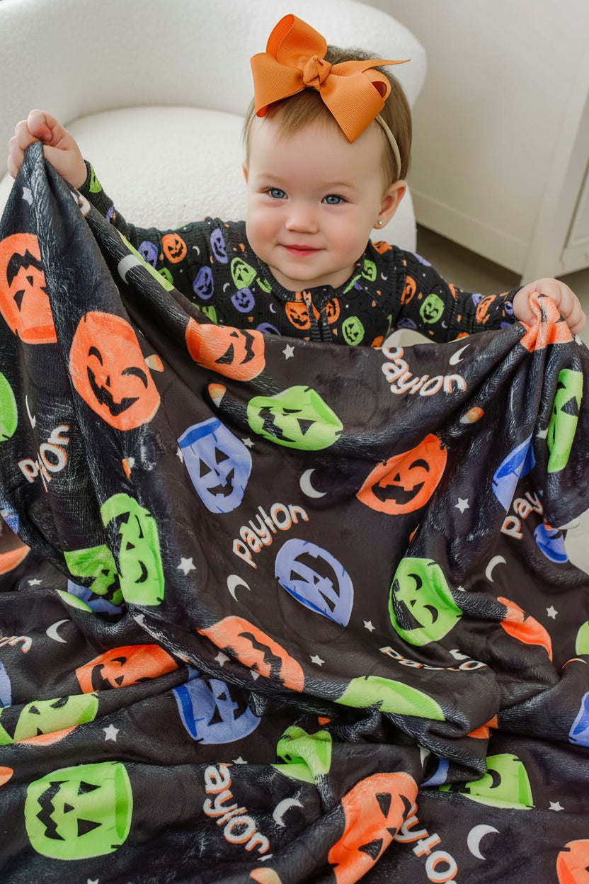 Baby with an orange bow holding up a personalized Halloween blanket with colorful jack-o-lantern print and repeating name text
