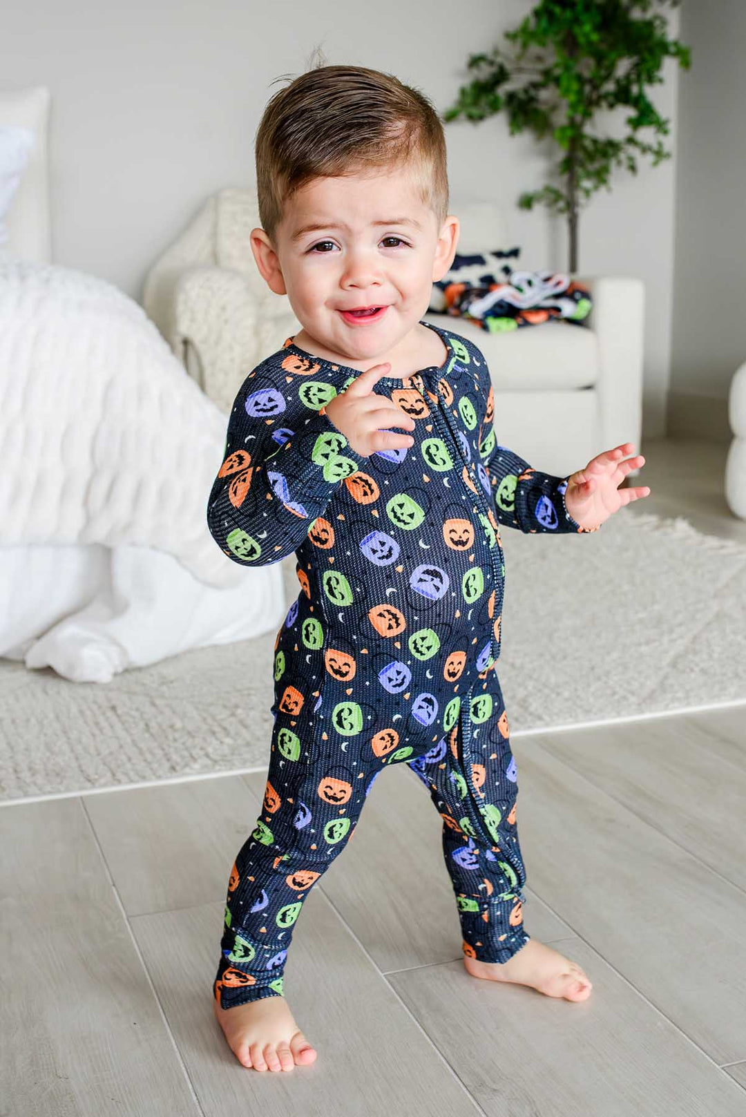 Toddler smiling and dancing in a living room wearing a bamboo waffle romper with colorful jack-o-lantern print