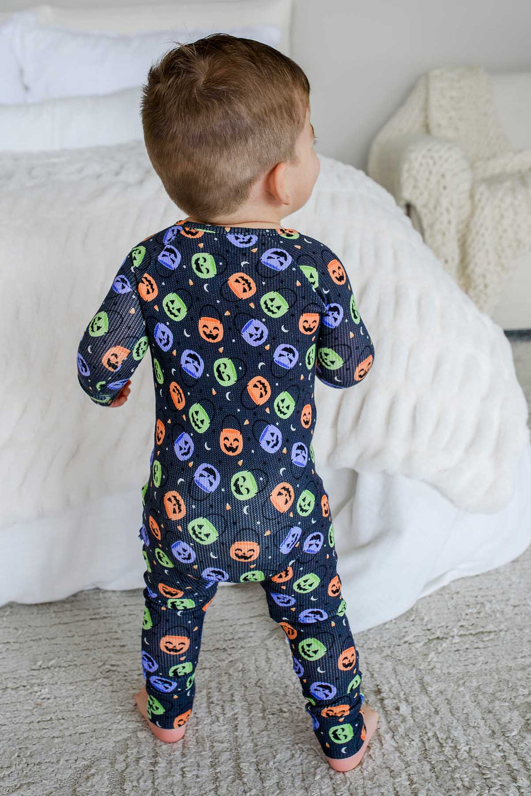 Back view of a toddler standing beside a bed in a bamboo waffle romper with colorful jack-o-lantern print