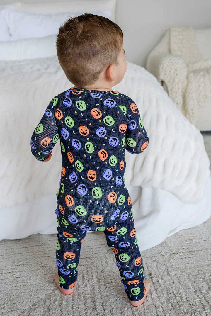 Back view of a toddler standing beside a bed in a bamboo waffle romper with colorful jack-o-lantern print