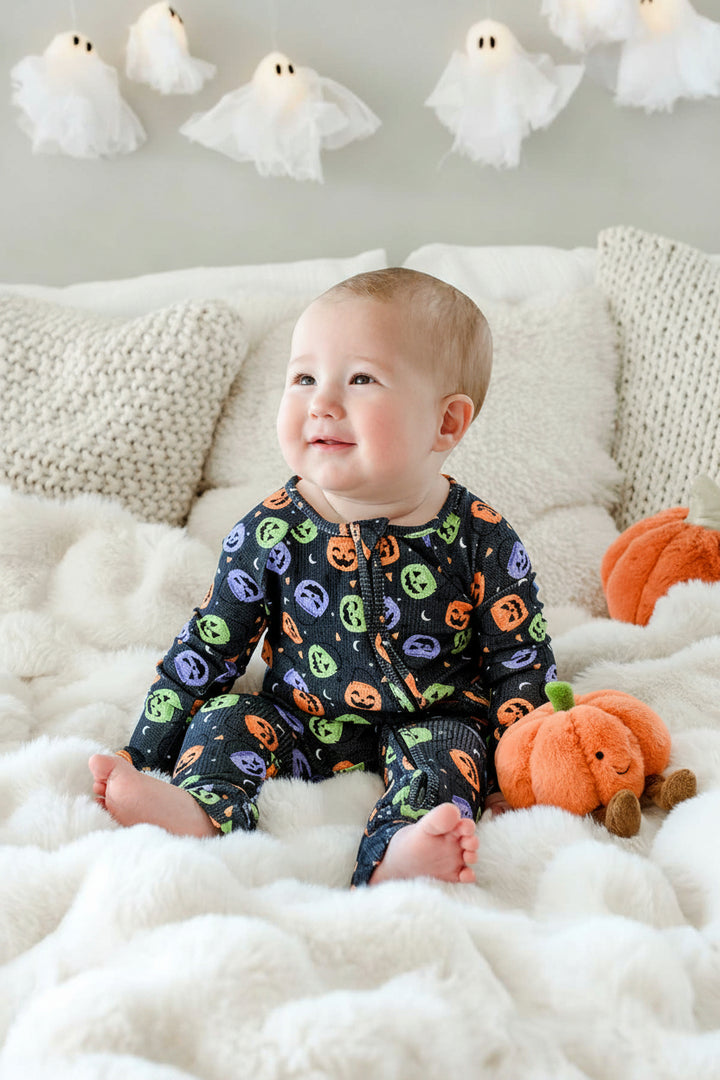 Baby sitting on a bed in a bamboo waffle romper with colorful jack-o-lantern print, beside a plush pumpkin toy, ghost lights hanging on wall behind