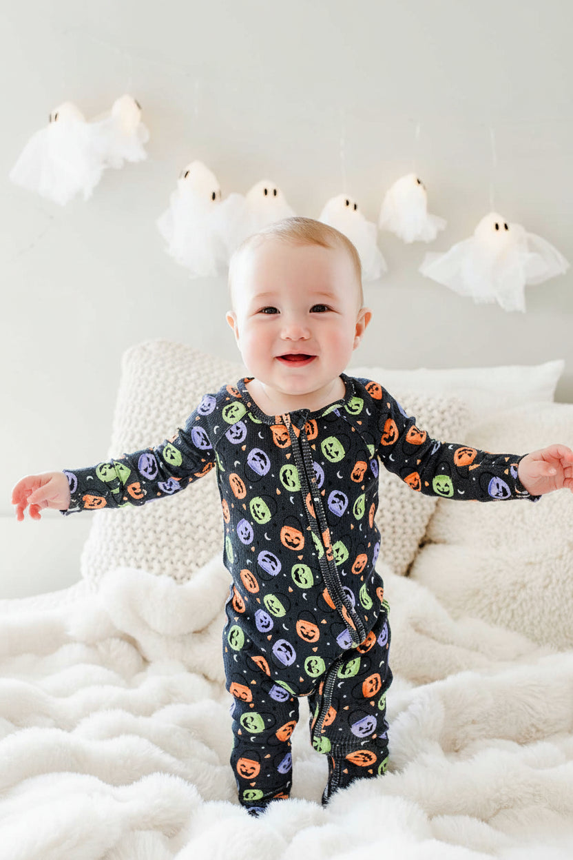 Baby standing on a bed smiling in a bamboo waffle romper with colorful jack-o-lantern print, ghost lights hanging on wall behind