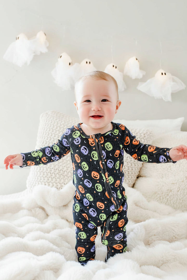 Baby standing on a bed smiling in a bamboo waffle romper with colorful jack-o-lantern print, ghost lights hanging on wall behind