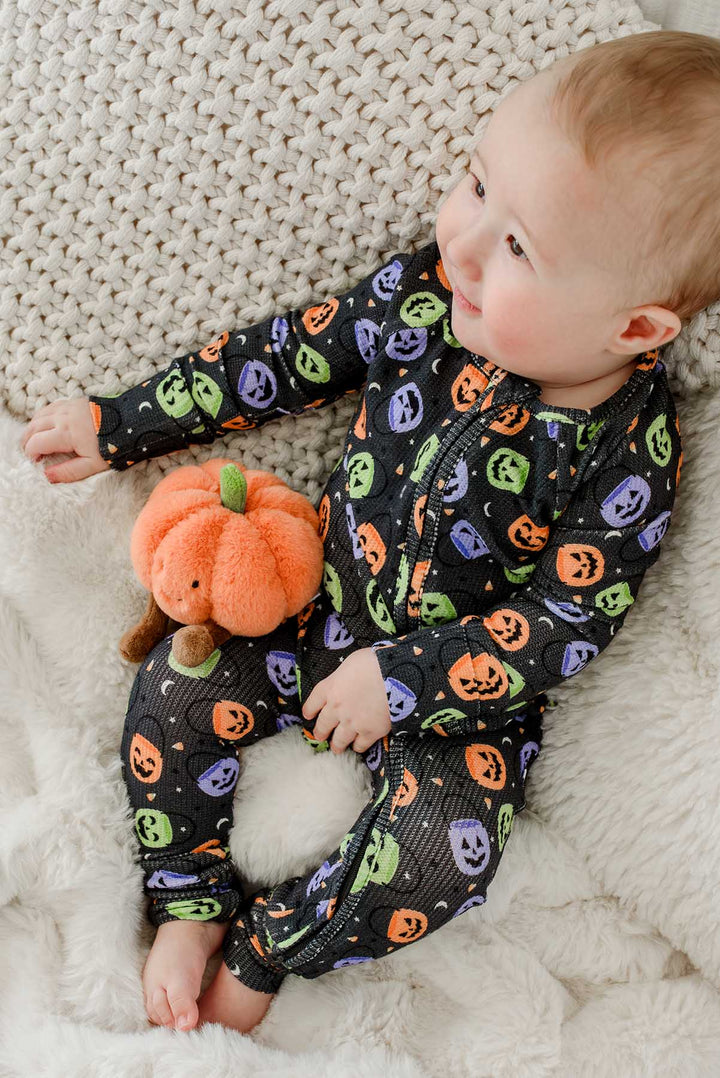 Baby sitting on a blanket in a bamboo waffle romper with colorful jack-o-lantern print, holding a plush pumpkin toy