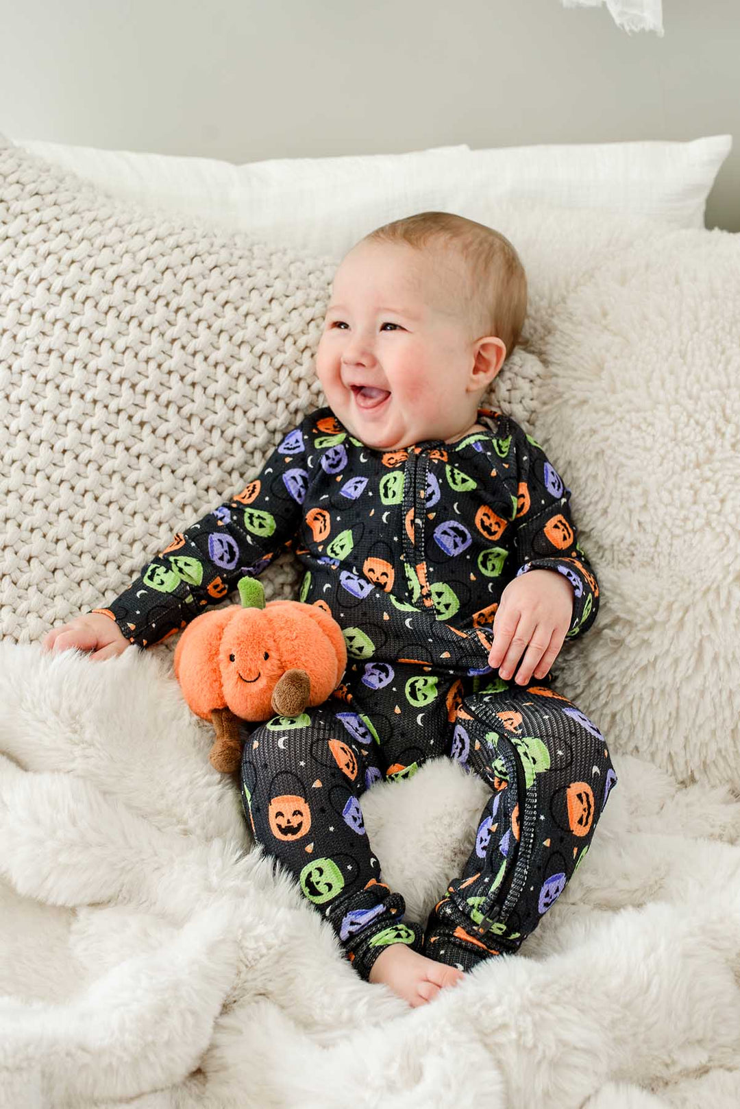Laughing baby sitting on a bed in a bamboo waffle romper with colorful jack-o-lantern print, holding a plush pumpkin toy