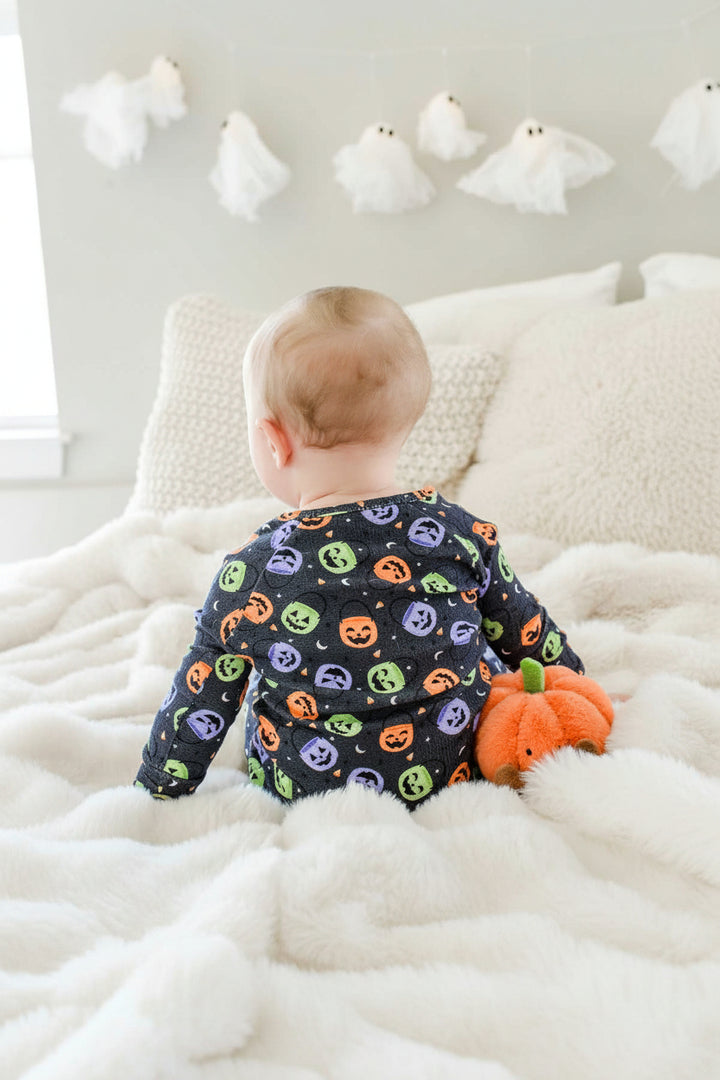 Baby sitting on a bed in a bamboo waffle romper with colorful jack-o-lantern print, back view, with a plush pumpkin toy and ghost decorations on wall