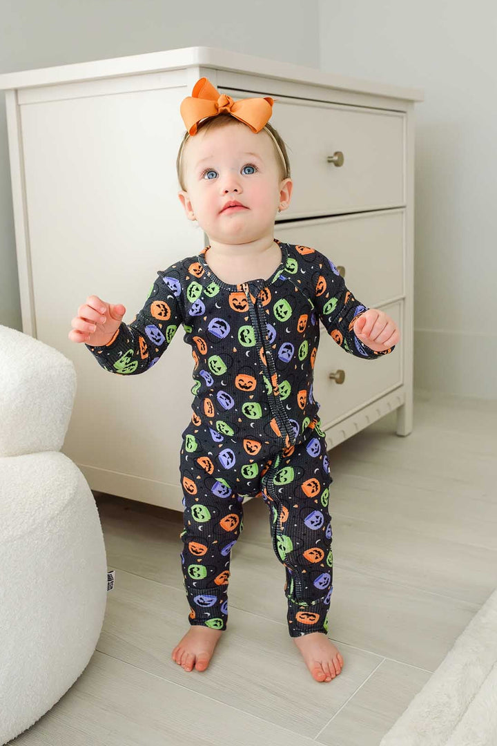 Toddler with an orange bow standing in a bamboo waffle romper with colorful jack-o-lantern print
