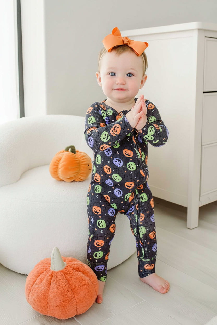 Toddler with an orange bow standing in a bamboo waffle convertible zip romper with colorful jack-o-lantern print, beside plush pumpkin decor