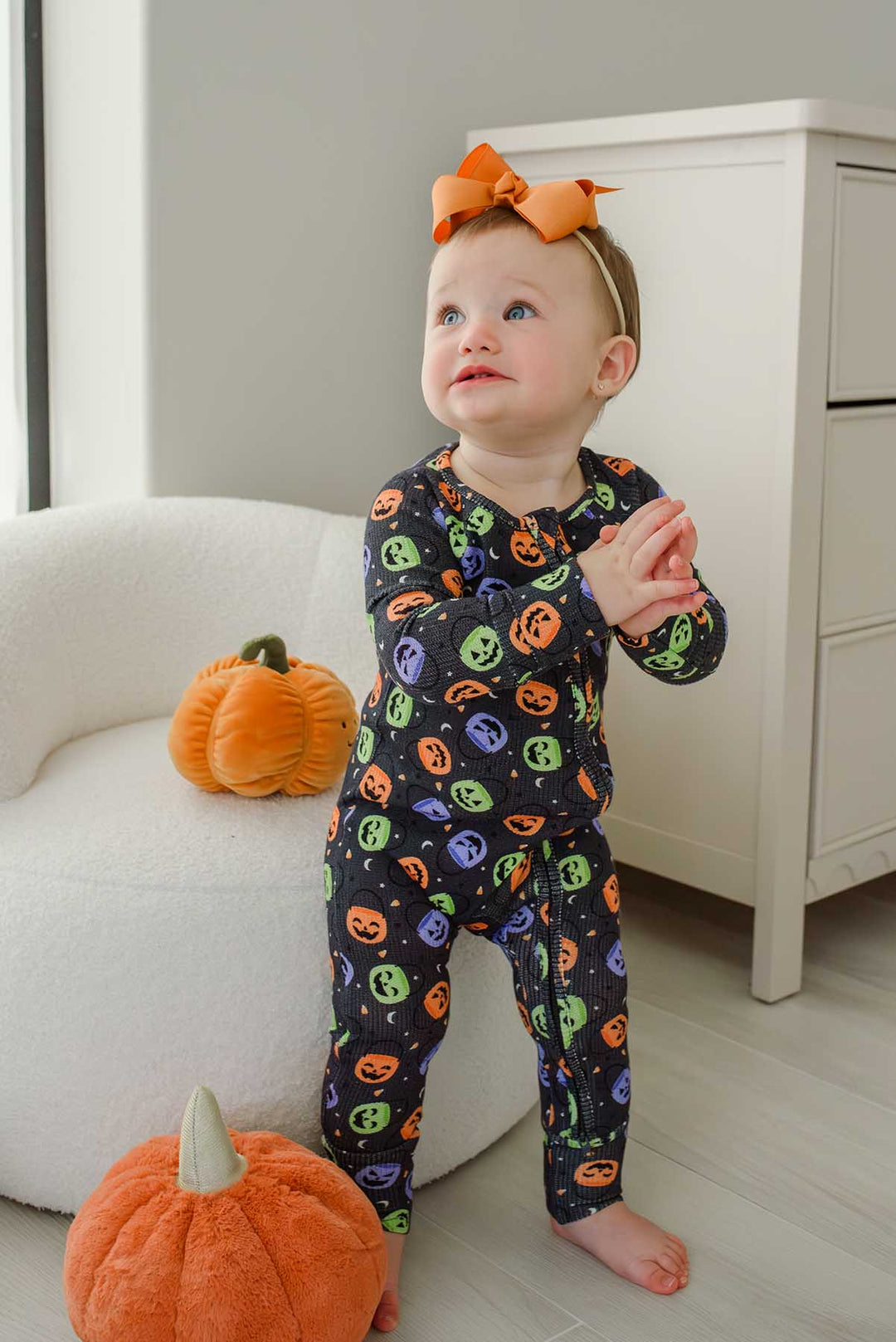 Baby with an orange bow clapping, standing beside plush pumpkin decor, wearing a bamboo waffle romper with colorful jack-o-lantern print