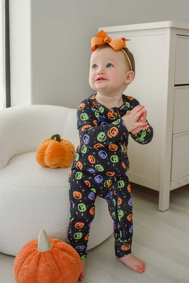 Baby with an orange bow clapping, standing beside plush pumpkin decor, wearing a bamboo waffle romper with colorful jack-o-lantern print
