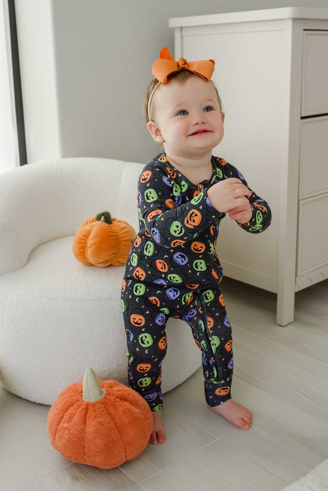Baby with an orange bow standing beside plush pumpkin decor, wearing a bamboo waffle romper with colorful jack-o-lantern print
