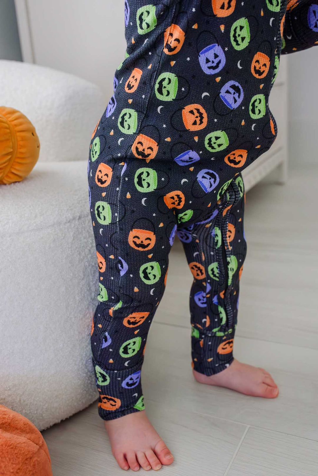 Close-up of a toddler's legs in a bamboo waffle romper with colorful jack-o-lantern print, standing beside a plush pumpkin