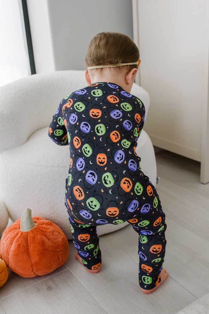 Back view of a baby climbing on furniture beside a plush pumpkin, wearing a bamboo waffle romper with colorful jack-o-lantern print