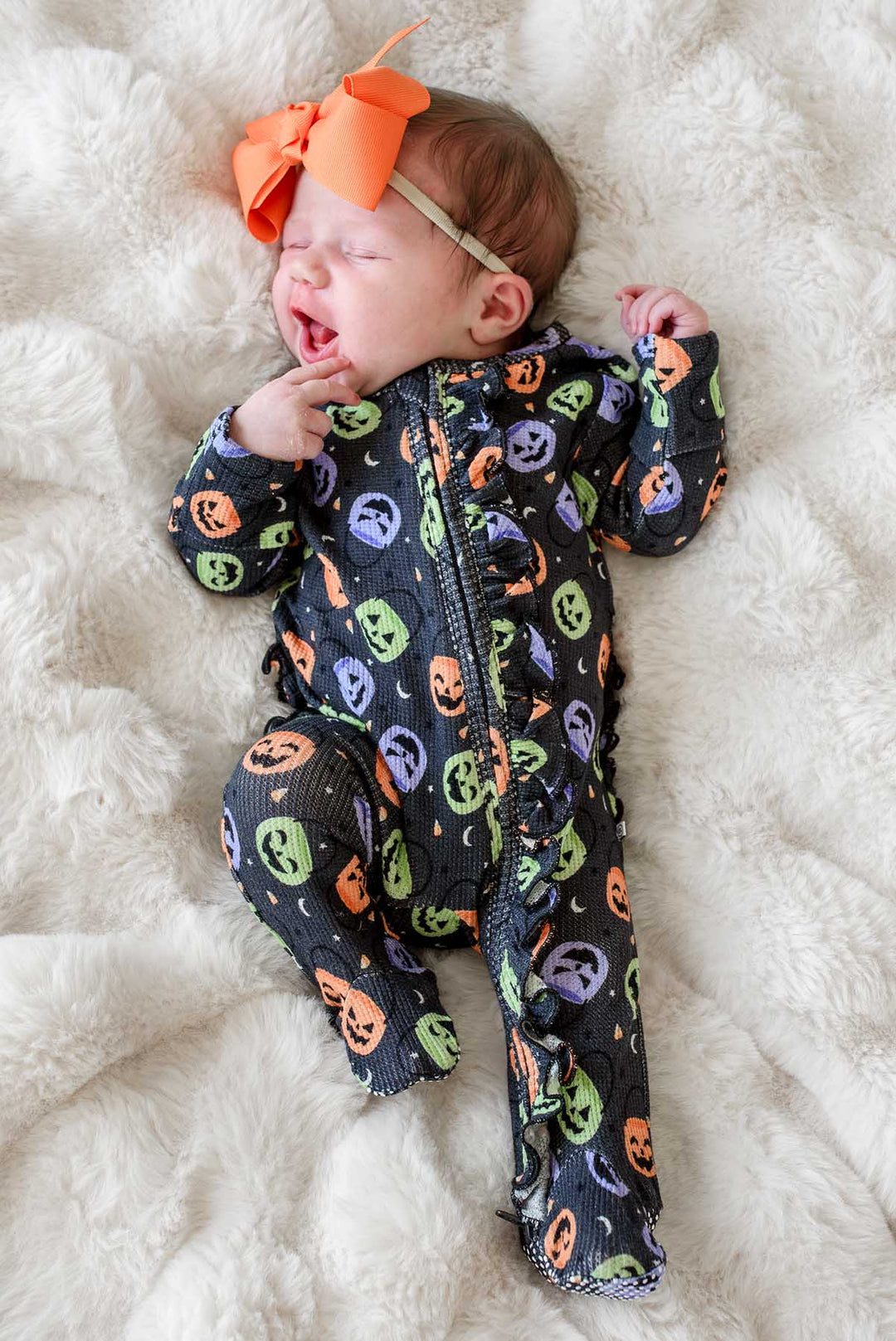 Yawning newborn with an orange bow wearing a bamboo waffle ruffle footie with colorful jack-o-lantern print, lying on a fluffy blanket