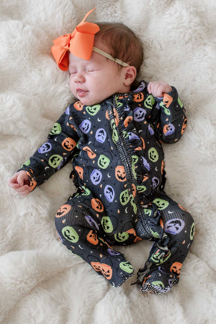 Sleeping baby with an orange bow wearing a bamboo waffle ruffle footie with colorful jack-o-lantern print, lying on a fluffy blanket