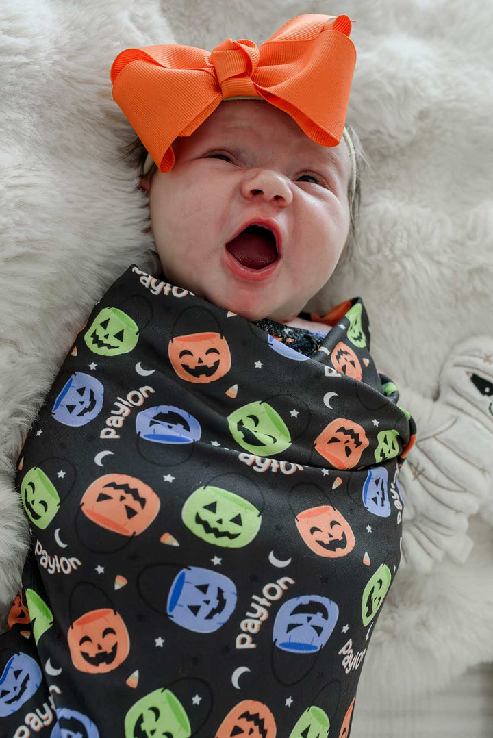 Yawning newborn wearing a large orange bow, swaddled in a personalized Halloween blanket with colorful jack-o-lantern print and name text