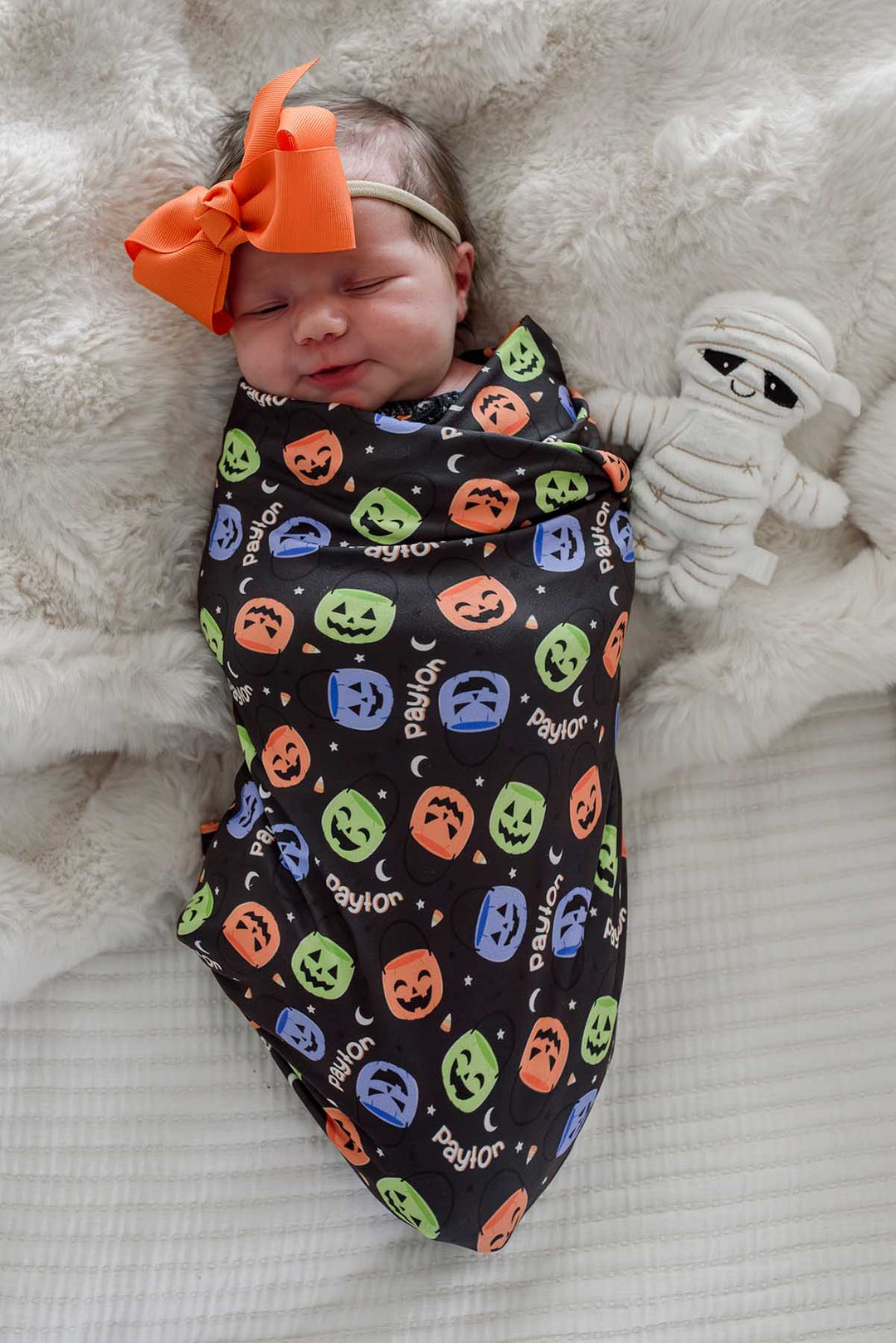 Sleeping newborn wearing an orange bow, swaddled in a personalized Halloween blanket with colorful jack-o-lantern print, beside a mummy plush toy