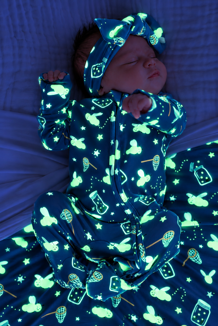 Overhead shot of a newborn stretching, wearing a Caden Lane blue bamboo zipper footie in a Chasing Fireflies print featuring fireflies and mason jars, paired with a bow headwrap.