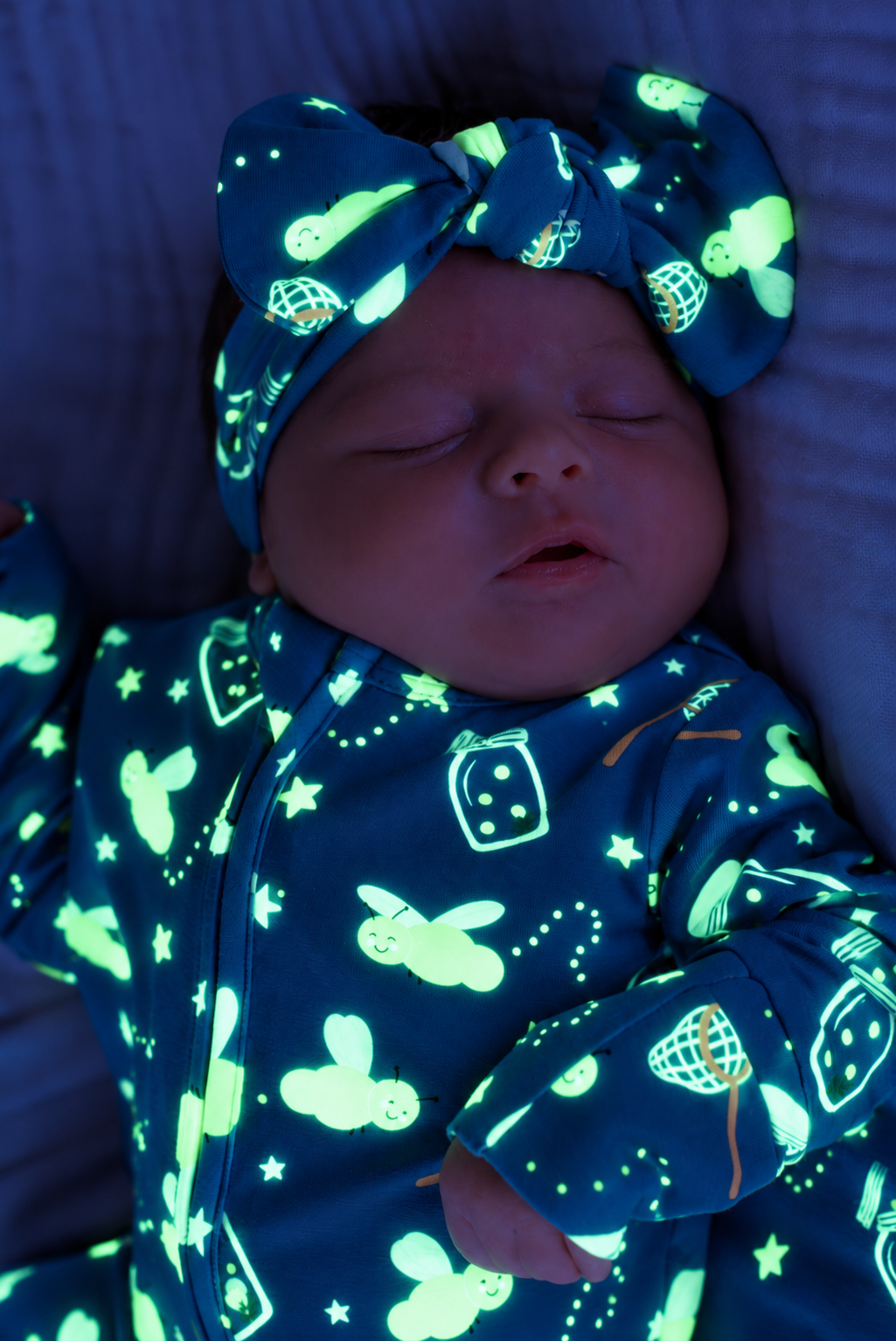 Close-up of a sleeping newborn wearing a Caden Lane blue glow-in-the-dark knit bow headwrap in a Chasing Fireflies print featuring fireflies and mason jars, paired with a matching footie.